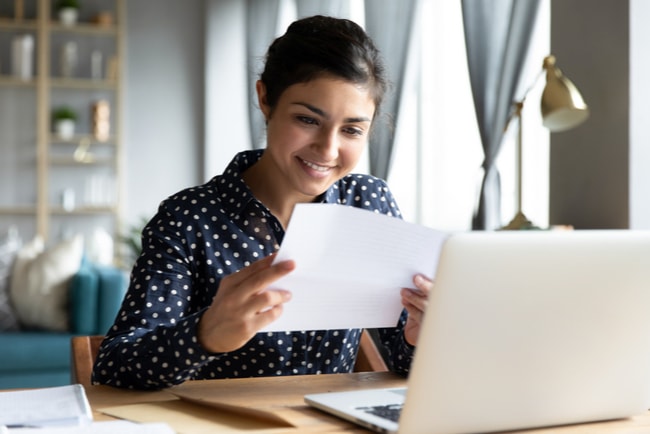 Ung kvinna sitter vid ett bord med en laptop och håller upp ett papper framför sig och ler.